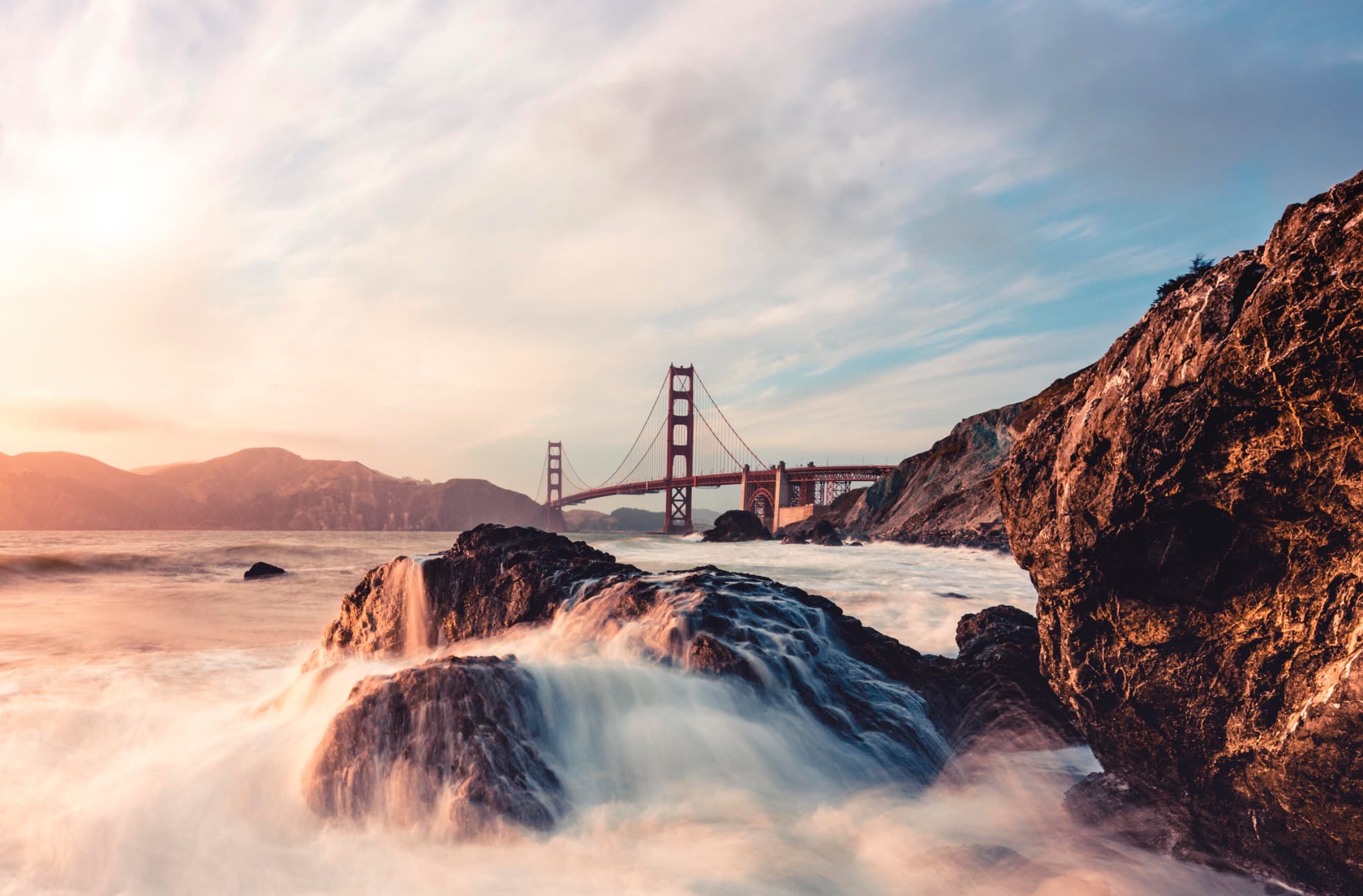 Waves washing over dark rocks on the San Francisco shoreline with the Golden Gate Bridge in the background at sunset